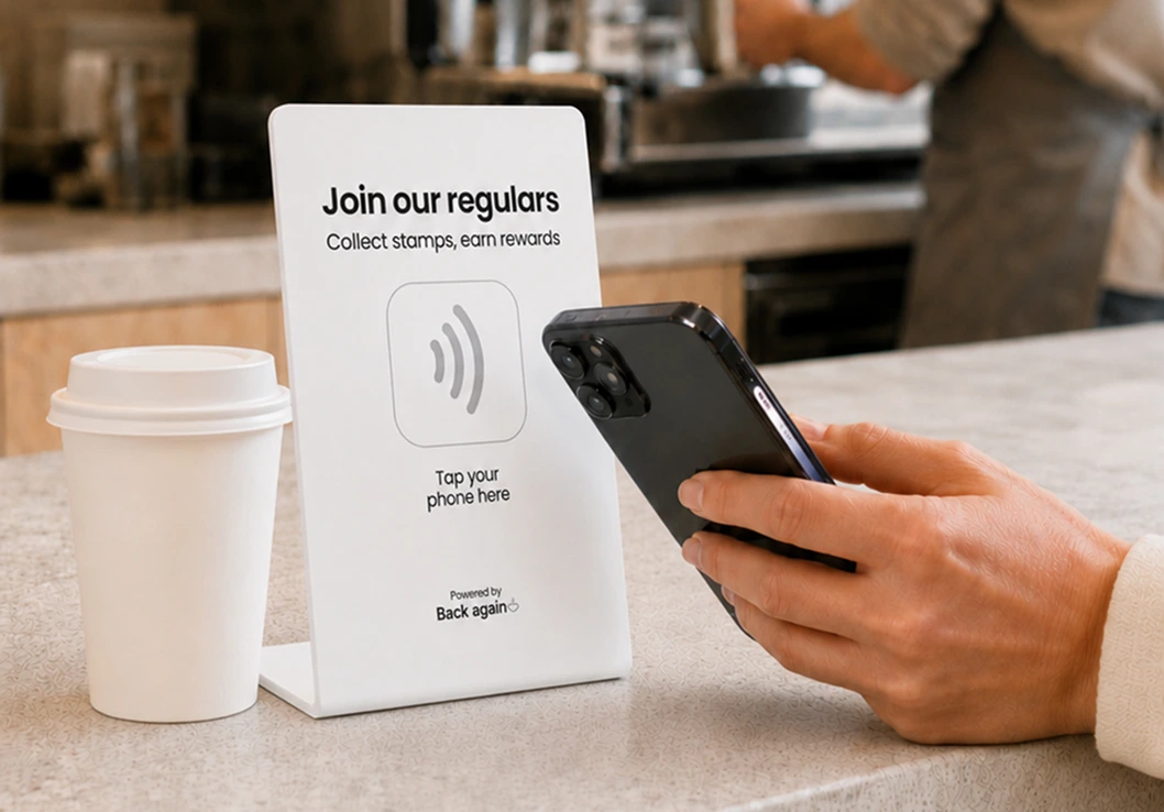 A customer tapping their phone on a Back Again loyalty sign at a cafe counter to collect a stamp